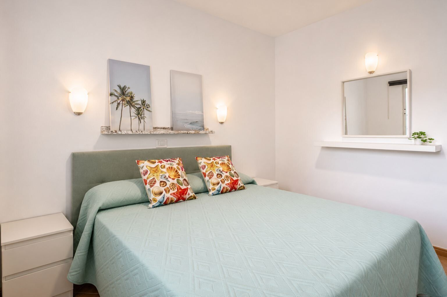A bedroom with a bed, pillows, a nightstand, a shelf with decorative items, and wall-mounted lights.