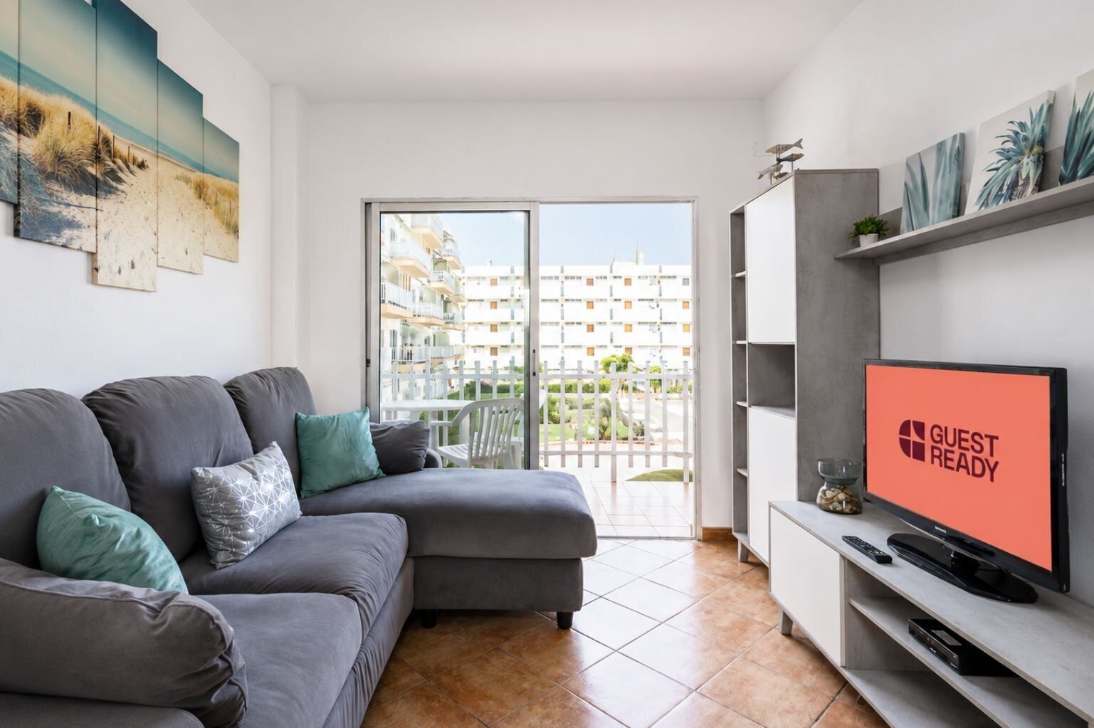 A modern living room with a grey sofa, a flat-screen TV, and a balcony with a view of buildings.