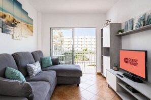 A modern living room with a grey sofa, a flat-screen TV, and a balcony with a view of buildings. - GuestReady - Tenesor Sun Beach Resort (Las Palmas de Gran Canaria)