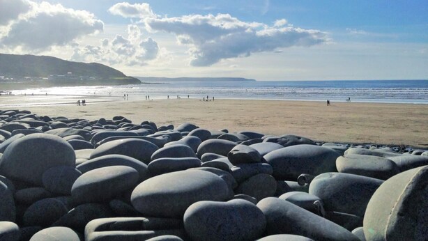 Ein Strand mit großen, glatten Steinen im Vordergrund, ein klarer Himmel und das Meer im Hintergrund.