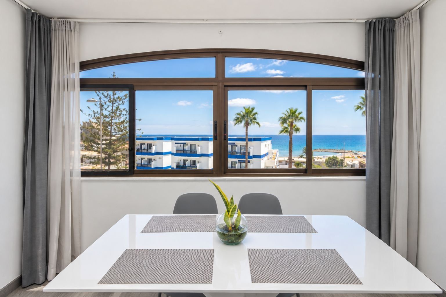 Ein Konferenzraum mit Blick auf einen Strand und Gebäude, ausgestattet mit einem weißen Tisch, zwei Unterlagen und einem Blumentopf.