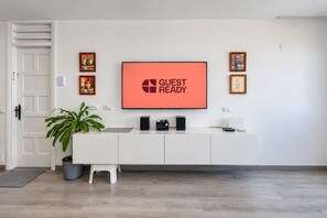 A modern living room with a flat-screen TV displaying 'GUEST READY,' a white console table with electronic devices, a potted plant, and framed artwork on the wall. - GuestReady - San Agustin Blue Flag Beach (San Agustin)