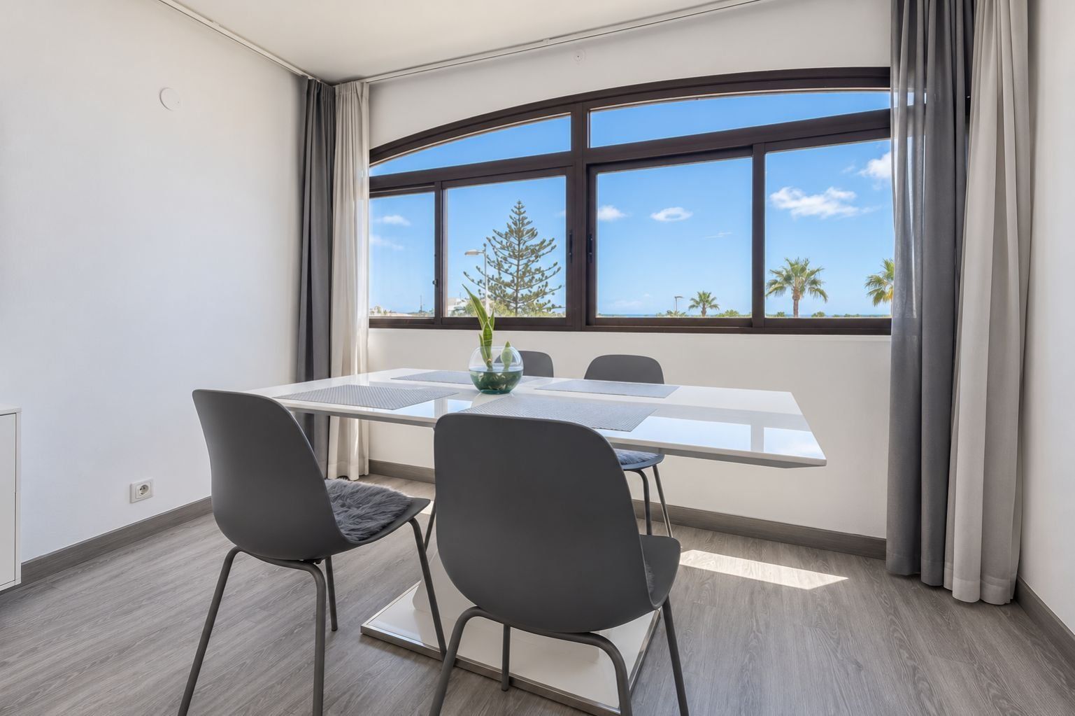 Una habitación moderna con un ventanal grande, una mesa blanca con dos sillas y una planta en maceta.