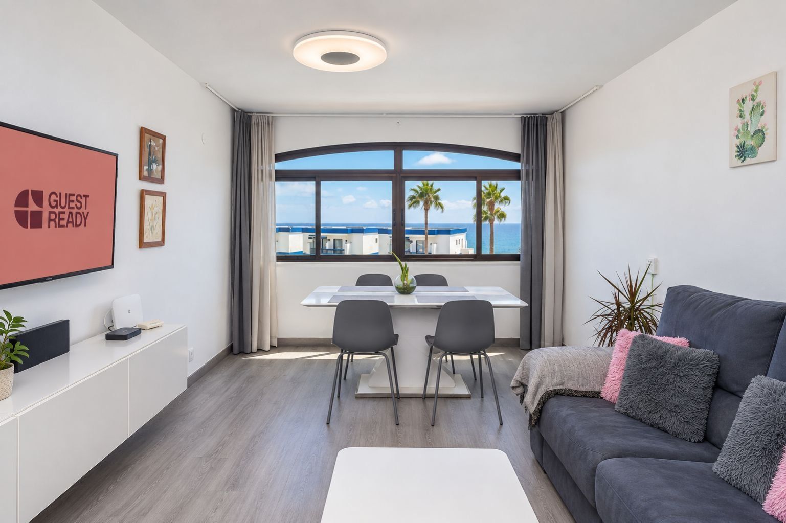 A modern hotel room with a dining area, a sofa, and a large window with a view of palm trees and the sea.