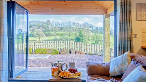 Una stanza accogliente con soffitto in legno, un divano in pelle marrone, un tavolo con prodotti per la colazione e vista su un paesaggio rurale attraverso una porta in vetro.