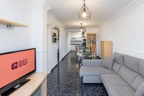 Ein modernes Wohnzimmer mit Flachbildfernseher, einem grauen Sofa und einem Essbereich mit Glastisch und Stühlen.