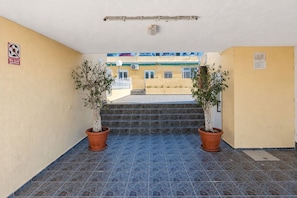 Ein Gebäudeeingang mit Fliesenboden, zwei Blumentöpfen, einem Schild mit der Aufschrift 'Kein Ballspielen' und Blick auf ein Gebäude mit blauen Fensterläden.