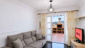 Sala de estar com sofá bege, televisão e vista para um pátio com churrasqueira.
