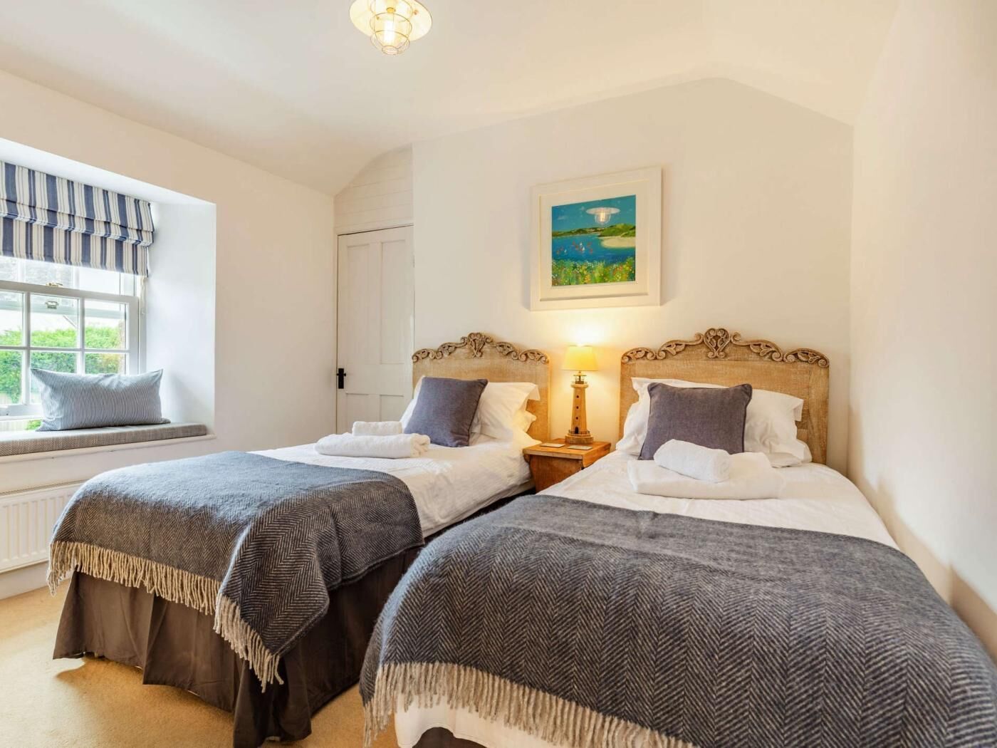 A room with two beds, a window with blinds, a painting on the wall, and a lamp on the nightstand.
