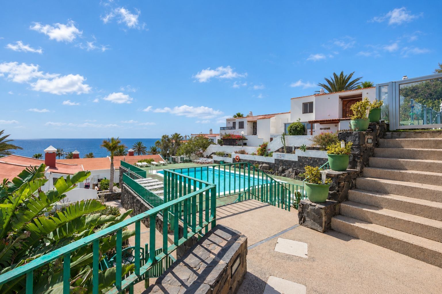 Zona de piscina con vista despejada al mar, una escalera que lleva a un edificio y plantas en macetas.