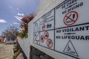 Um aviso com múltiplas advertências e regras para uma área de natação, incluindo proibição de mergulho, ausência de salva-vidas e proibição de nadar com crianças menores de 12 anos sem a supervisão de um adulto.