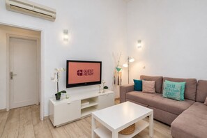 Ein modernes Wohnzimmer mit einem Flachbildfernseher, der 'GUEST READY' anzeigt, einem Sofa mit türkisfarbenen und beigen Kissen, einem weißen Couchtisch und einer weißen TV-Konsole mit einer kleinen Pflanze und einer Kerze.