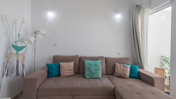 A modern living room with a brown sectional sofa, teal and beige cushions, a dreamcatcher, and a large window with curtains. - GuestReady - Aguila Beachfront Apartment (San Agustin)