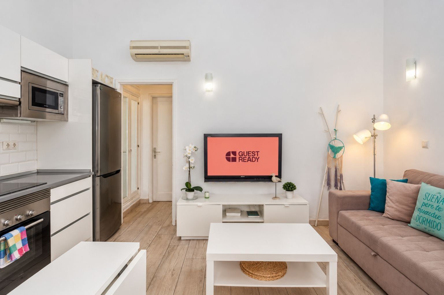 A modern kitchen with a microwave, oven, and refrigerator. There's a flat-screen TV on a white stand with a 'GUEST READY' sign. A white sofa with teal pillows is in the living area.