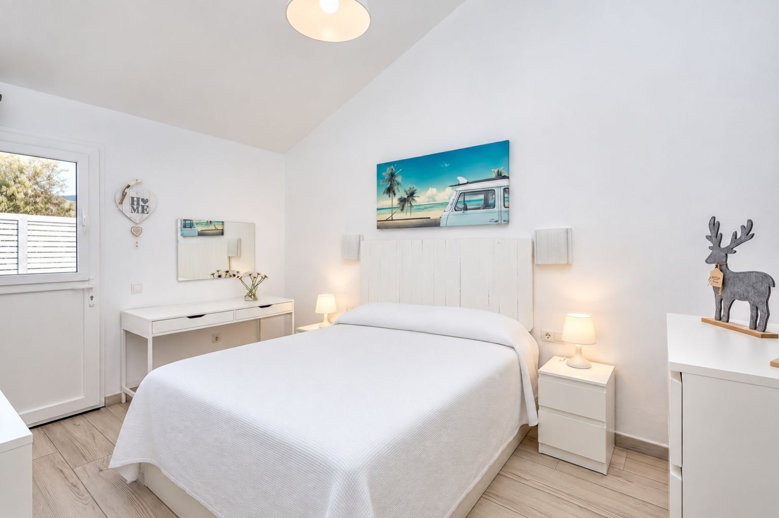 Ein Hotelzimmer mit einem Bett, Nachttischen, einem Schminktisch, einem Fenster mit Jalousien, einem Bild an der Wand und einem dekorativen Rentier.