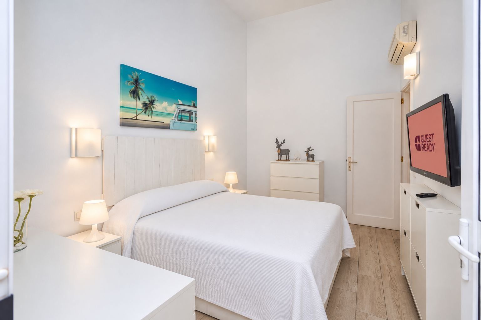 Une chambre d’hôtel avec un lit, des tables de chevet, un téléviseur affichant le message « CHAMBRE PRÊTE POUR L’ARRIVÉE DES CLIENTS », un tableau représentant une scène de plage et une porte.