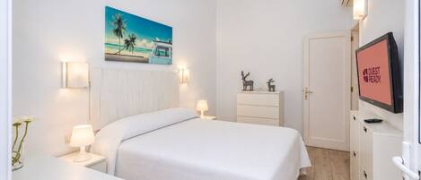 A hotel room with a bed, bedside tables, a TV displaying 'GUEST READY', a painting of a beach scene, and a door.
