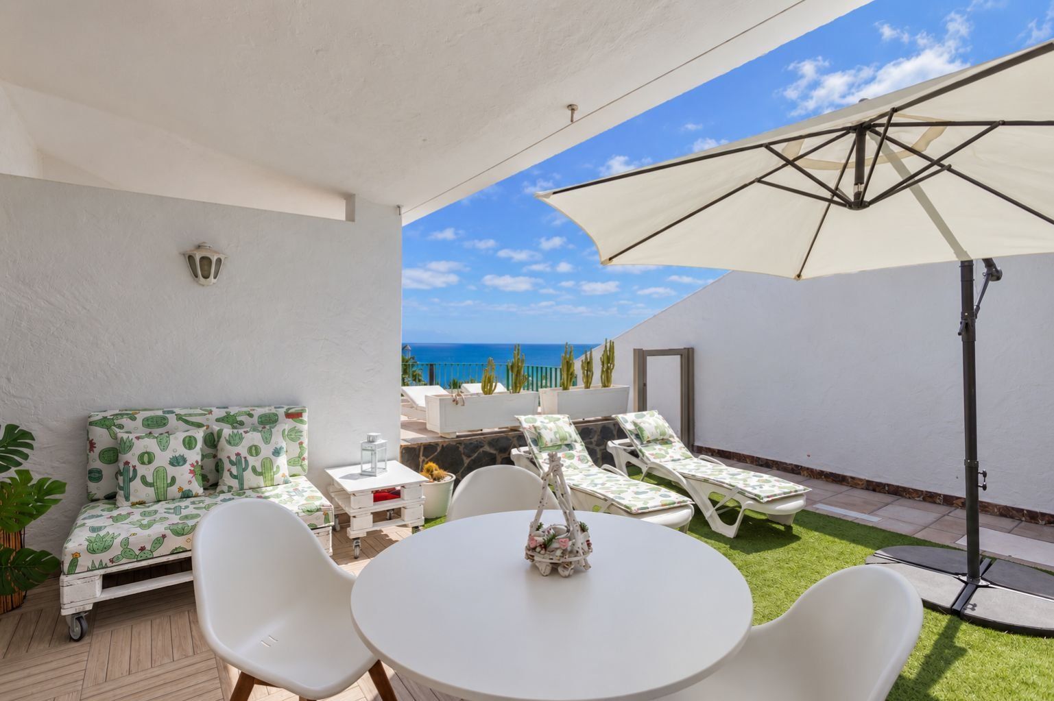 Un patio con una mesa y sillas blancas, un sofá blanco con estampados de cactus y un paraguas grande.