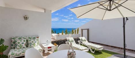 Un patio con una mesa y sillas blancas, un sofá blanco con estampados de cactus y un paraguas grande.