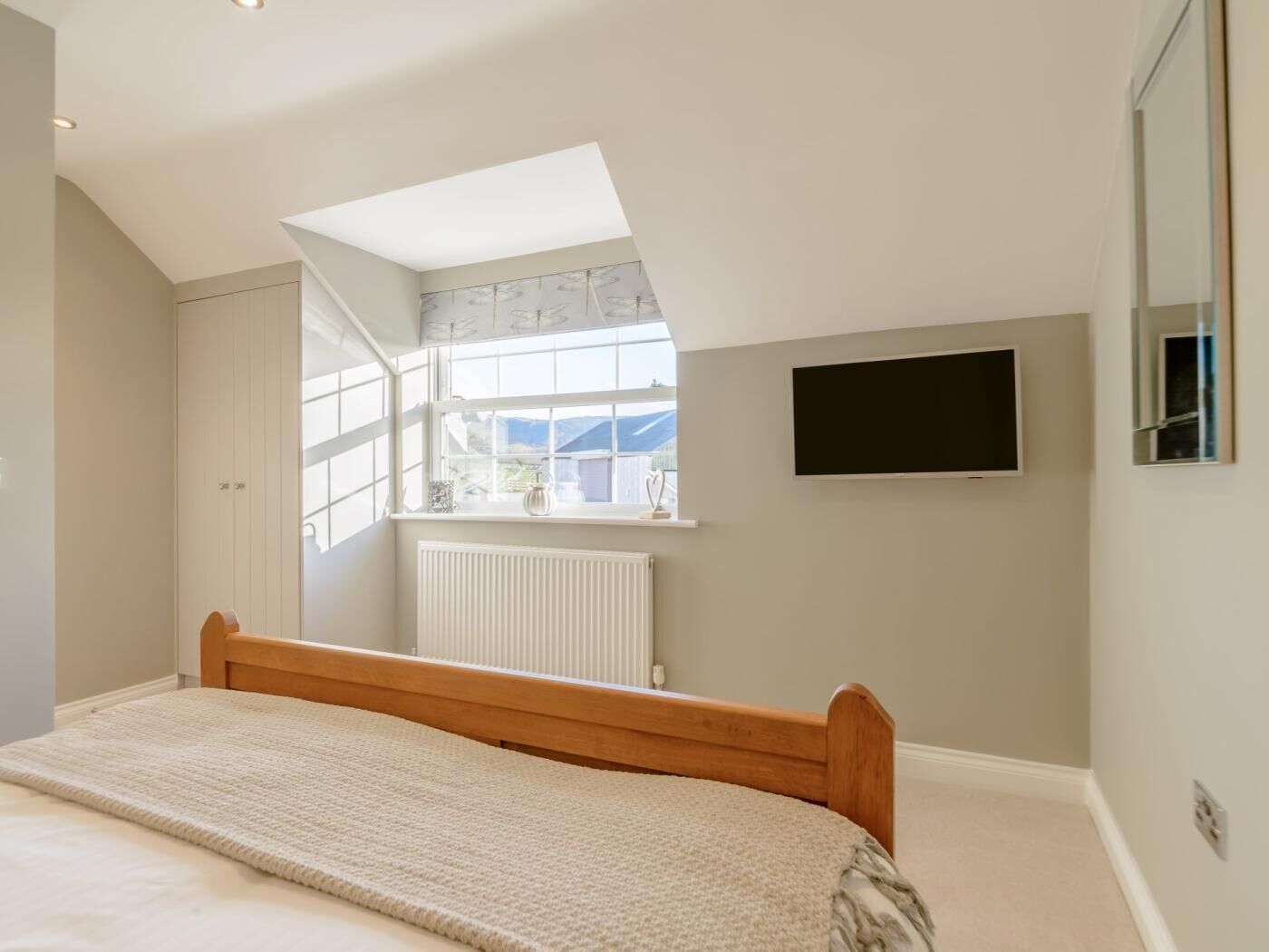Een slaapkamer met schuine balken, een bed, een raam met uitzicht, een radiator en een aan de muur bevestigde televisie.