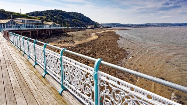 Eine hölzerne Pier mit aufwendigen weißen Geländern, die sich in einen Strandbereich mit sanften Hügeln und blauem Himmel erstreckt.