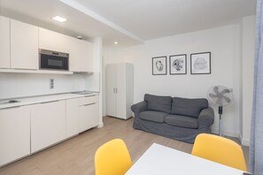 Eine moderne Küche mit weißen Schränken, einer Mikrowelle und einem Spülbecken. Im Wohnbereich befindet sich ein graues Sofa und ein gelber Sessel.