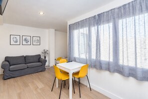 Ein modernes Wohnzimmer mit einem grauen Sofa, einem weißen Tisch und gelben Stühlen. An der Wand hängen gerahmte Bilder und ein großes Fenster mit transparenten Vorhängen.
