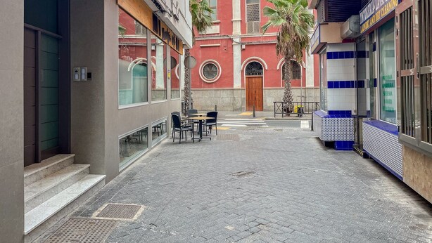 A narrow street with buildings on either side, a palm tree, and outdoor seating. - GuestReady - Artiles Near Canteras Beach (Las Palmas de Gran Canaria)