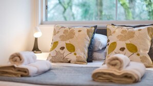 Ein ordentlich bezogenes Bett mit aufgerollten Handtüchern, zwei Kissen mit Blättermotiv und einer Lampe auf einem Nachttisch.
