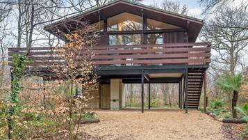 A modern two-story house with a large balcony, surrounded by trees and a gravel driveway.