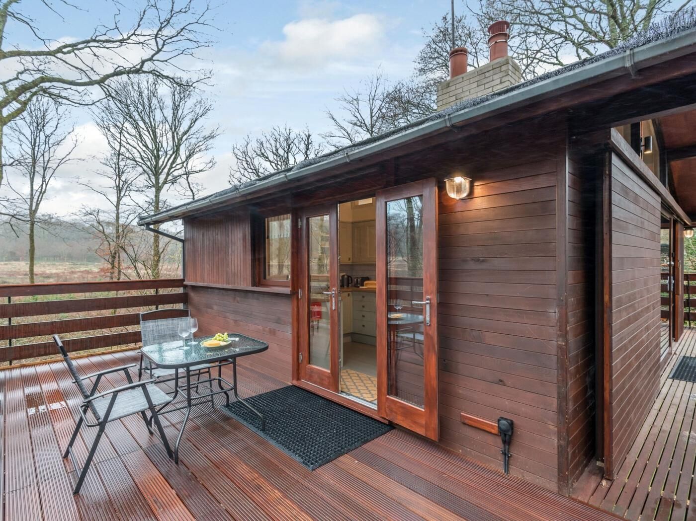 A wooden cabin with a deck, a glass door entrance, and a small outdoor table with a chair.