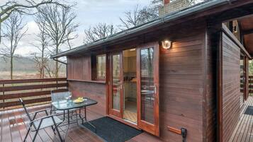 A wooden cabin with a deck, a glass door entrance, and a small outdoor table with a chair.