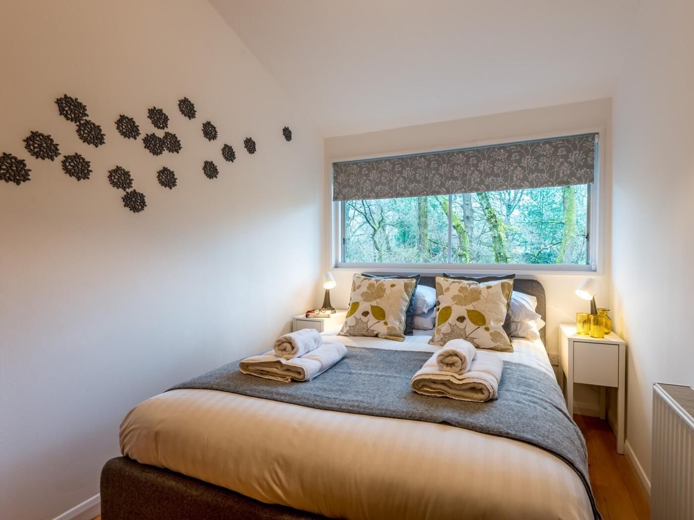Una cama bien hecha con un edredón gris y dos toallas enrolladas, una ventana con vistas a árboles y una pared con decoración.