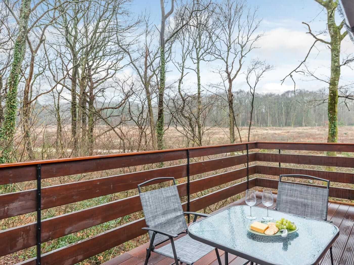 Una terraza de madera con una mesa y sillas de vidrio, con vistas a un área boscosa.
