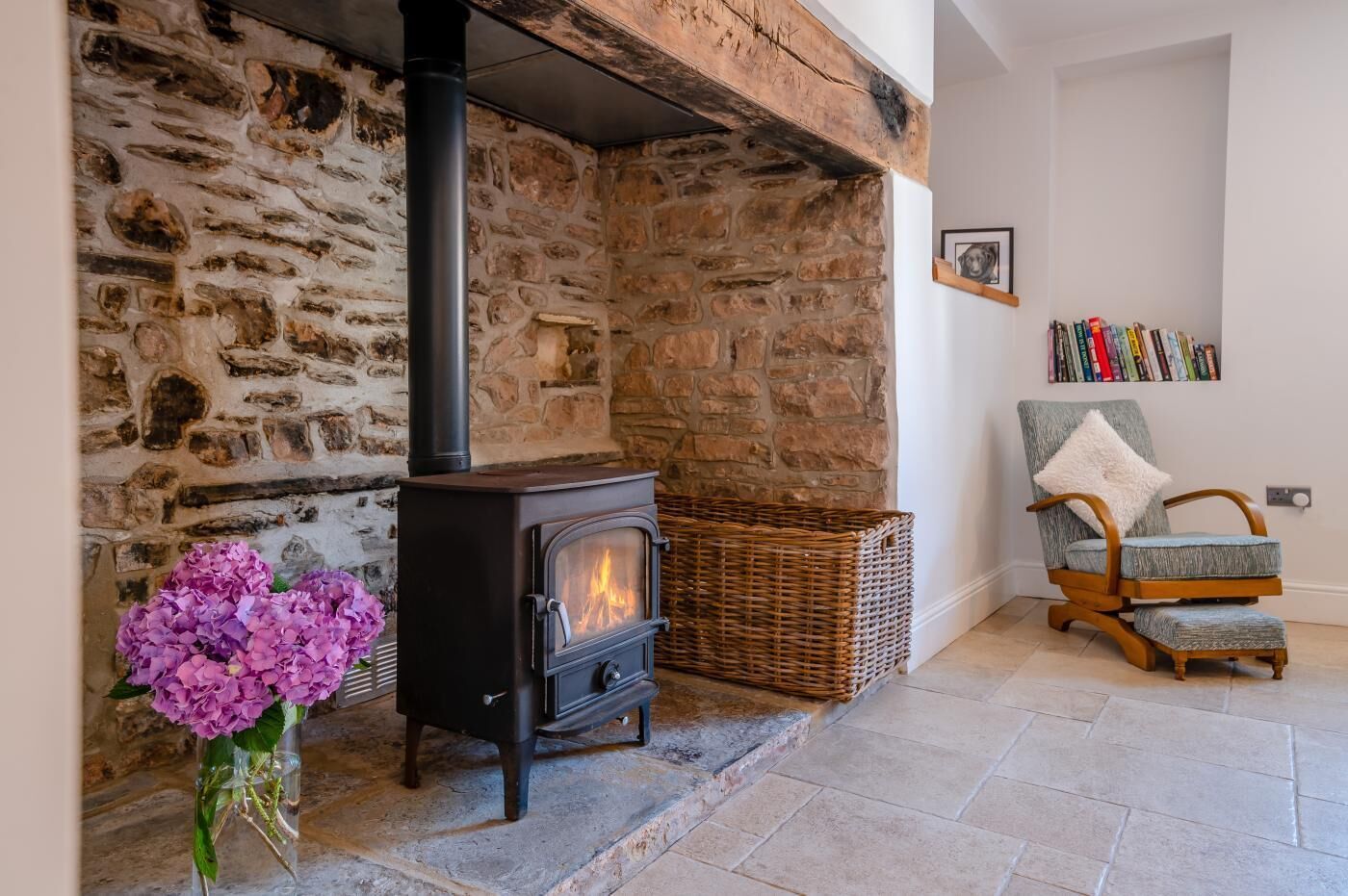 A stone fireplace with a black stove, a wicker basket, a chair with a fluffy pillow, and a vase of flowers.