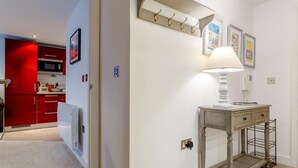 Ein Flur mit einer Lampe, einem Kleiderhaken und einem kleinen Tisch ebenfalls mit einer Lampe. Eine Tür führt in einen Küchenbereich mit roten Schränken und einer Mikrowelle.