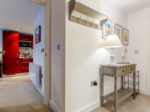 A hallway with a lamp, a coat rack, and a small table with a lamp. There is a door leading to a kitchen area with red cabinets and a microwave. - 1 Bed in Fowey (oc-77956) (Fowey)