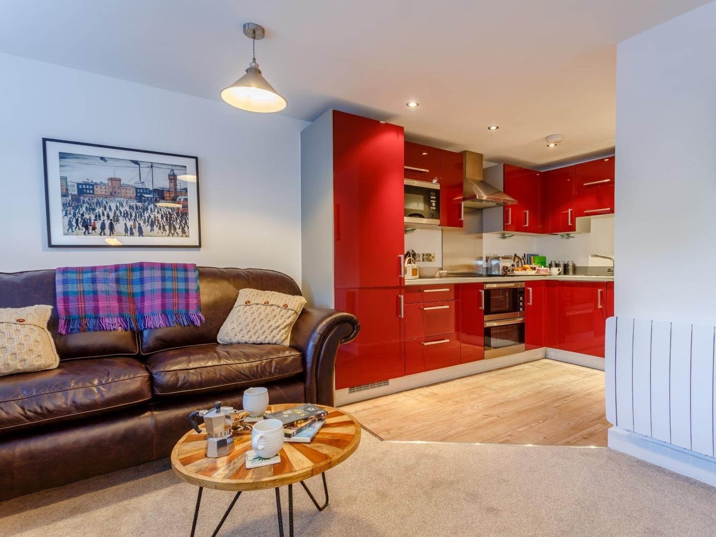 A living room with a brown leather sofa, a coffee table with a book and a coffee maker, and a red kitchen in the background.