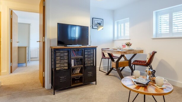 Ein Zimmer mit Fernseher auf einem Schrank, einem Esstisch mit Stühlen und einem Fenster mit Jalousien.