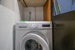 A compact, white washing machine in a small laundry room with a tiled wall and a wooden cabinet.