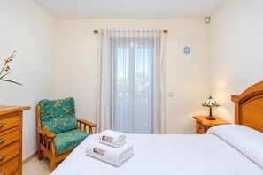 Une chambre d’hôtel comprenant un lit, une chaise, une table de chevet, une lampe, une fenêtre avec des rideaux et un thermostat fixé au mur.