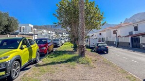 A street with parked cars, a palm tree, and buildings with white walls and red roofs.