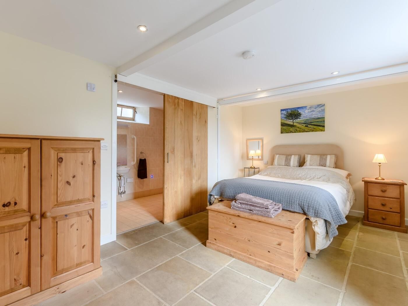 A bedroom with a bed, bedside tables, a wooden wardrobe, a bathroom with a sink, and a window.