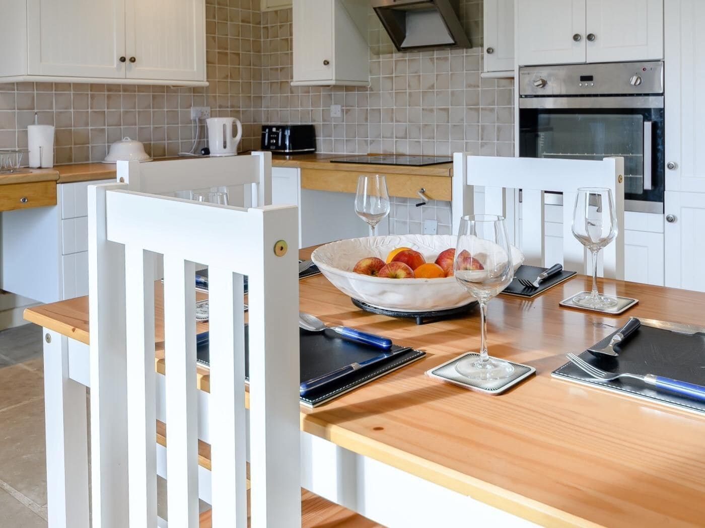 A modern kitchen with white cabinets, a wooden countertop, and a dining table set with a bowl of fruit, wine glasses, and cutlery.