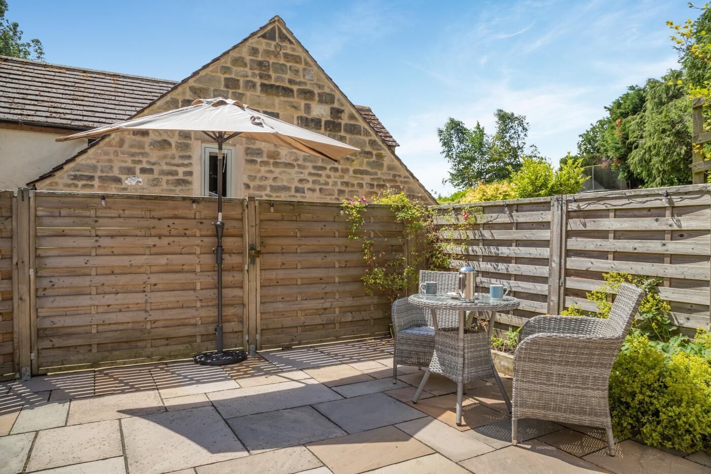 A stone building with a sloped roof, a wooden fence, and a patio area with wicker furniture and a table.
