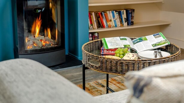 A cozy room with a fireplace, a wicker table with books and a magazine, and a bookshelf in the background. - 1 Bed in Matlock (oc-78225) (Paddocks)