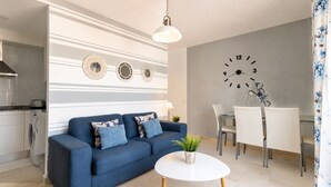 A modern living room with a sofa, a dining table with chairs, a clock on the wall, and a kitchen area with a washing machine. - GuestReady - Arenas Balcony and Pool (Caleta de Fuste)