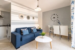 Ein modernes Wohnzimmer mit Sofa, einem Esstisch mit Stühlen, einer Wanduhr und einer Küchenzeile inklusive Waschmaschine.