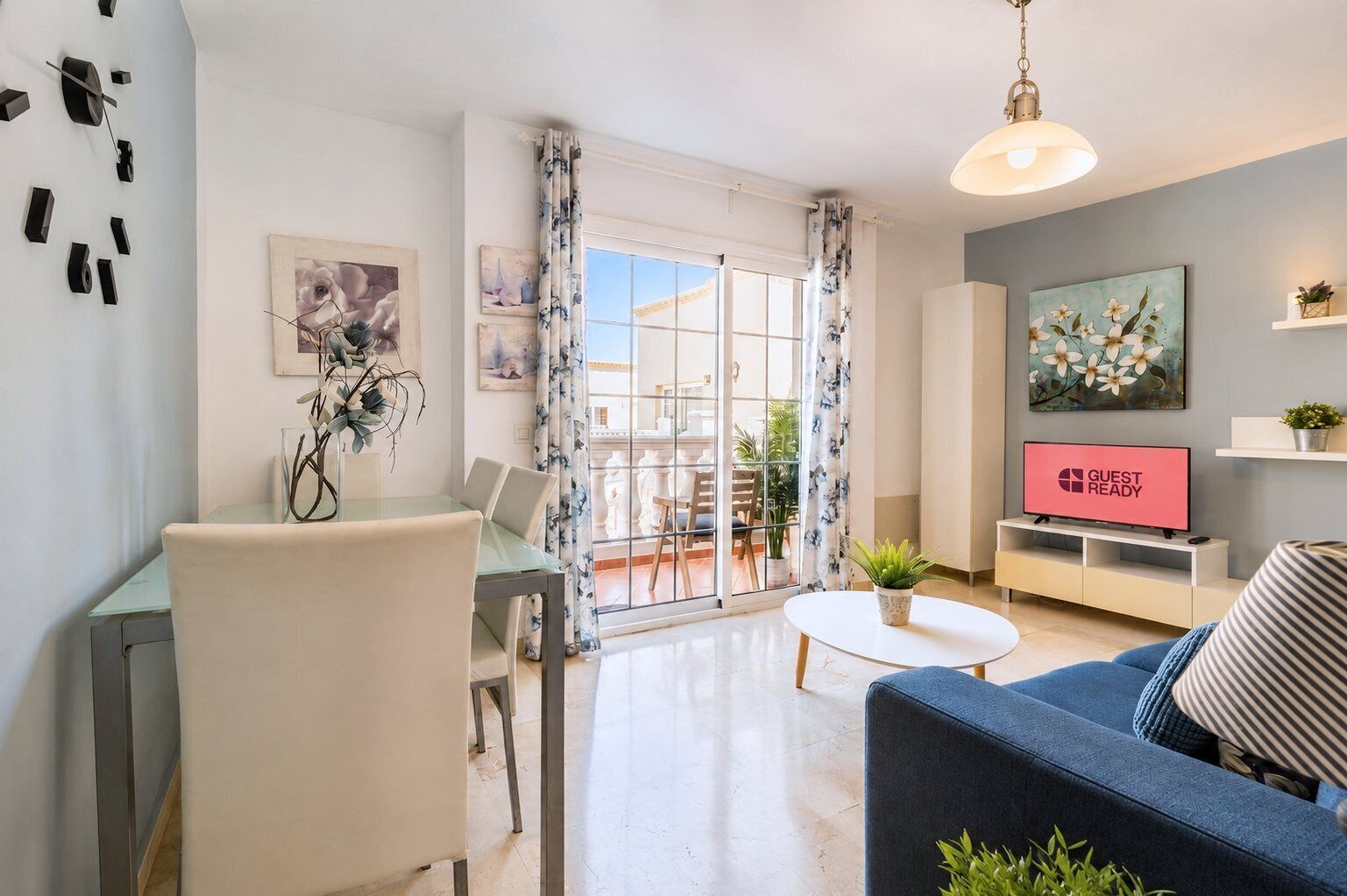 A modern living room with a sofa, a dining table with chairs, a TV displaying 'GUEST READY', and a balcony with a view.