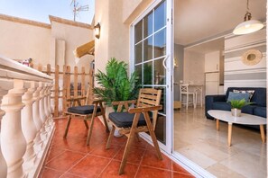 Ein Balkon mit Holzstühlen und einem Blumentopf, eine Schiebetür zu einem Innenraum mit einer Couch und einem Tisch.